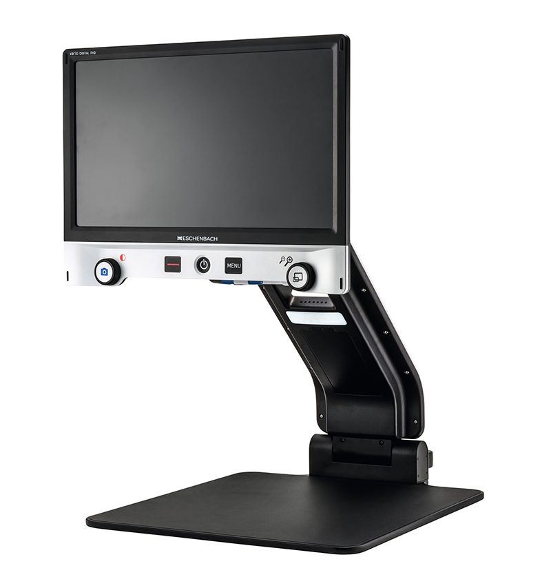 A Vario Digital FHD desktop video magnifier; a computer screen supported by a folding arm over a surface upon which items are placed, magnified, and displayed.